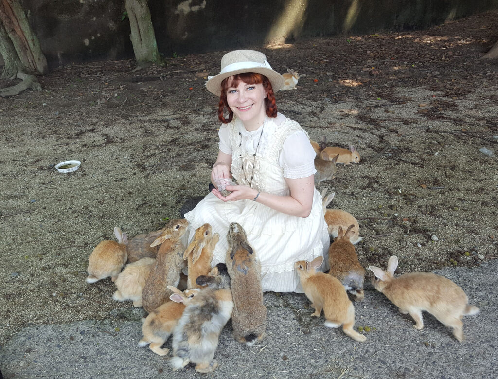 A white woman with red hair in braids, wearing a frilly white dress and straw hat, smiles at the camera from a crouch position. She is surrounded by about a dozen friendly, fluffy bunnies, some of whom are climbing onto her lap in search of the food she is holding.